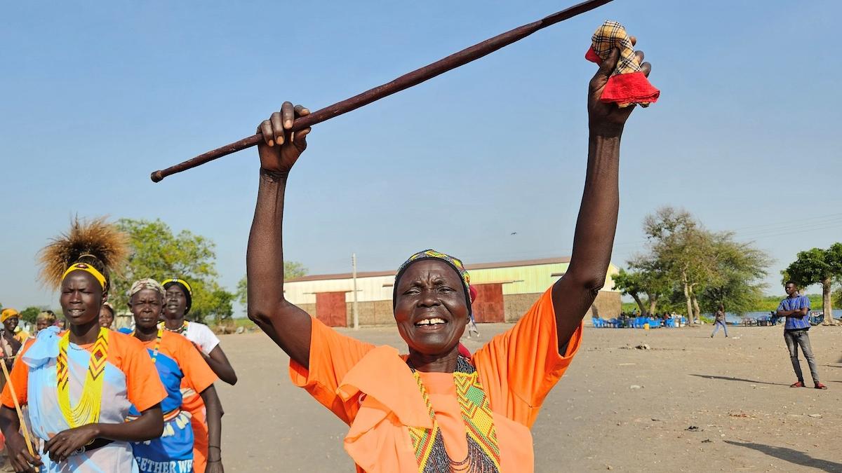 A woman wearing a bright orange t-shirt and large necklace is smiling and holding a wooden object above their head in jubilation. There are some other women in the background. 