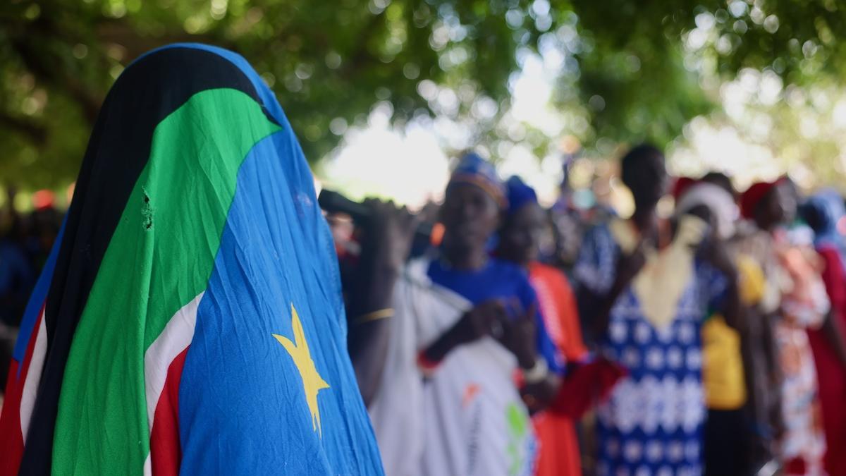 A person in the foreground is facing away from the camera and has covered their head with the South Sudanese flag. In the background, there are more people in bright clothing which have been blurred. All of the these people are standing under trees.