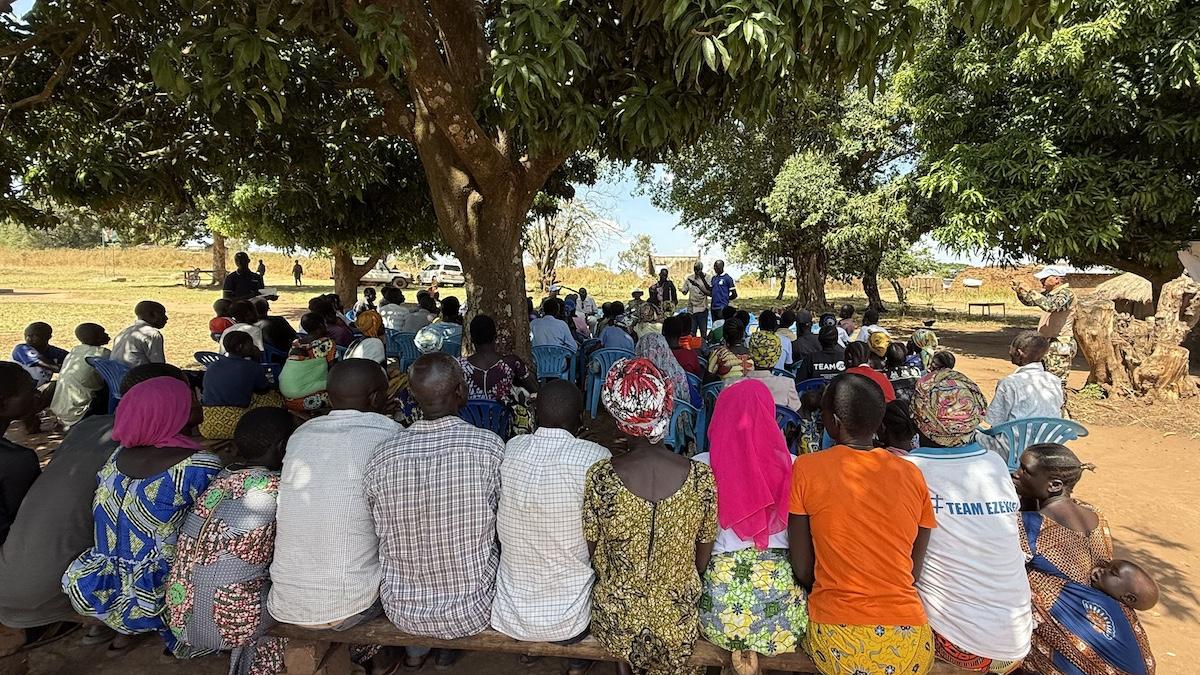 A group of people in brightly coloured clothing seated on wooden benches under a large tress are facing a person giving a presentation. 