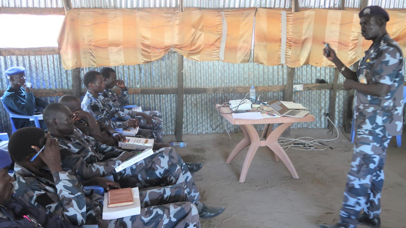 A group of uniformed police officers sitting watching a presentation being given by another uniformed person.