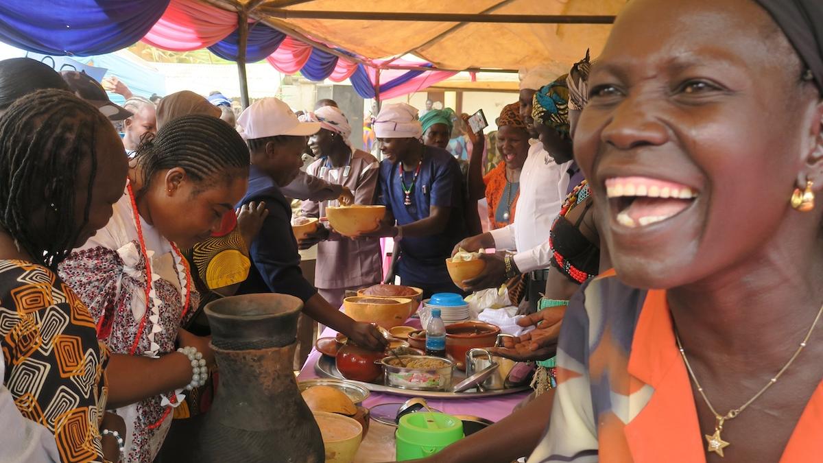 A woman is smiling as she sits in front of a table full of food. There are many other women sitting at the long table which is covered with a tent. 