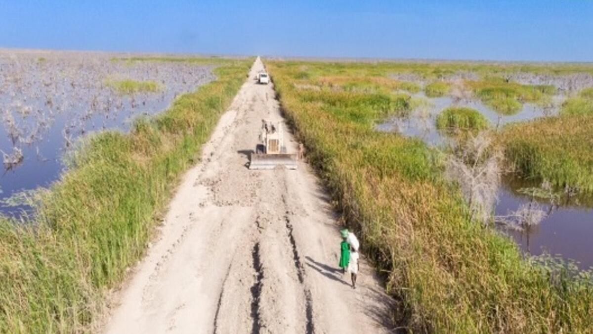 An aerial view of a dirt road with grass and bodies of water on either side. On the road, there are some people walking. 