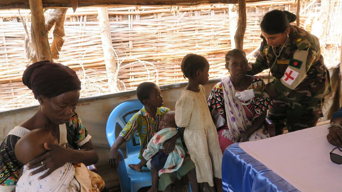 A uniformed medical officer is providing treatment to a seated woman. Next to the female patient is another woman and three children. 