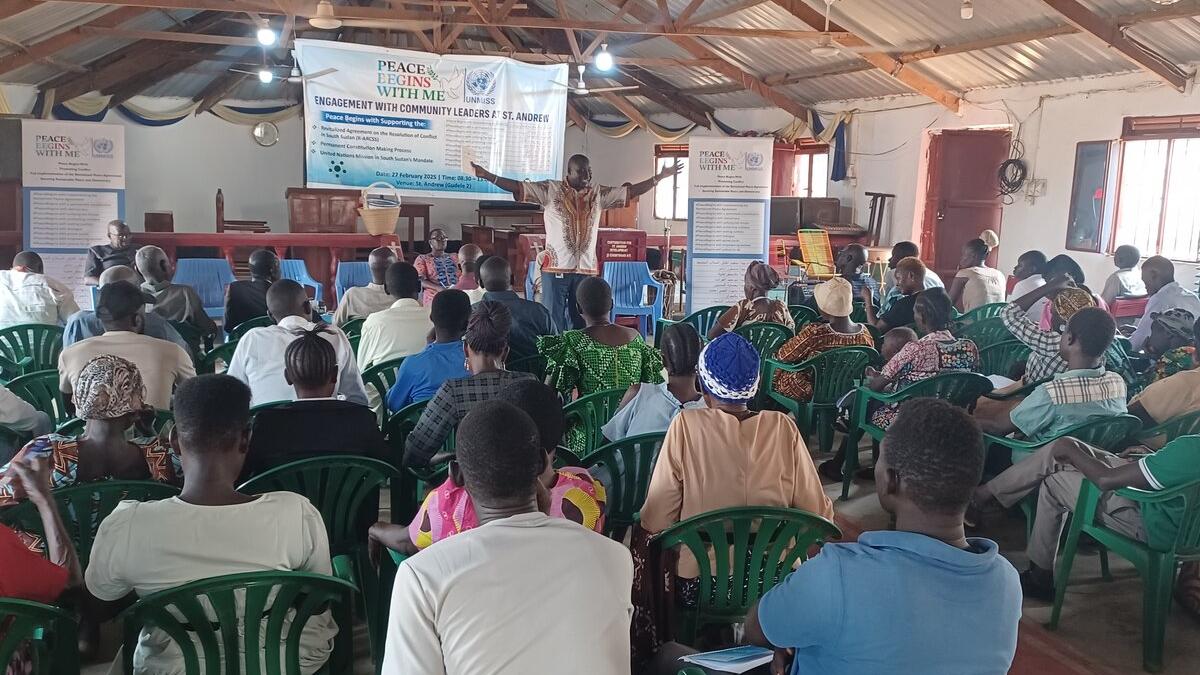 A group of people seated in green plastic chairs seated facing a stage where a man is presenting with their arms outstretched.