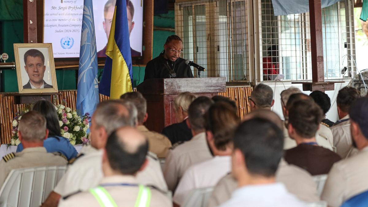 A group of people seated watching a person deliver an address at a podium in honour of a fallen peacekeeper.