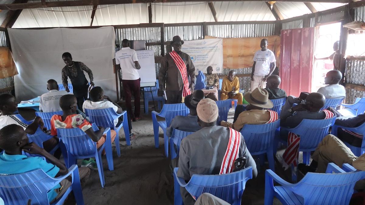 A group of people seated in blue chairs facing the front of the room where a presentation is being given.