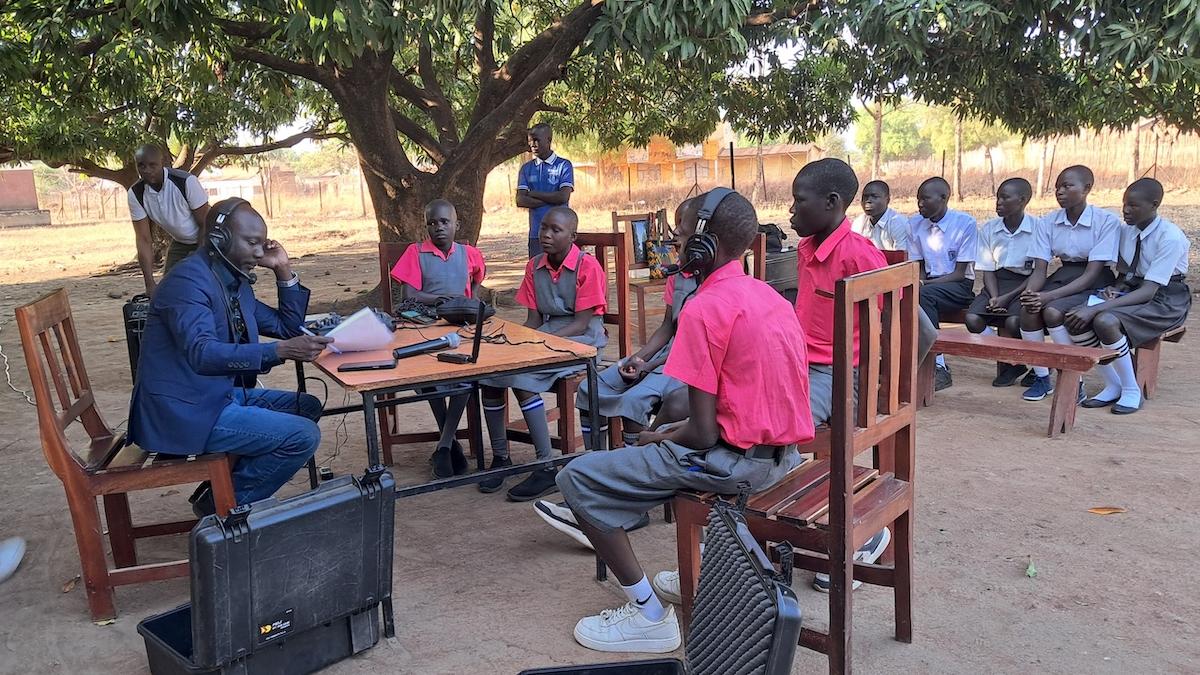 A man with a suit sits across a table with children in red t-shirts. All of the people at the table are wearing headphones and are recording a radio show. In the background, there are a group of students wearing blue shirts sitting watching the group at the table..