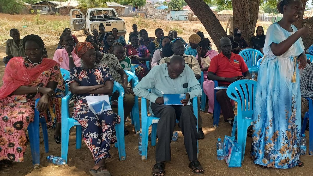 A group of people sitting in blue plastic chairs underneath a tree. In the background, there is a car with its door open.