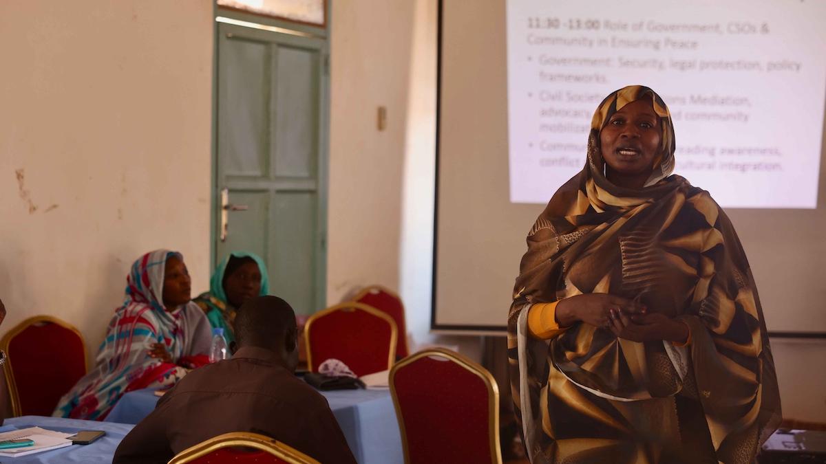 A woman addressing a group of people seated at tables. In the background, there is a PowerPoint presentation. that the woman is presenting.
