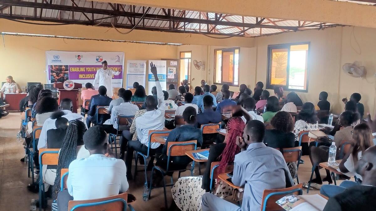 A classroom-style setting with many people seated at desks facing a speaker at the front, where banners and a presentation on youth empowerment are displayed.