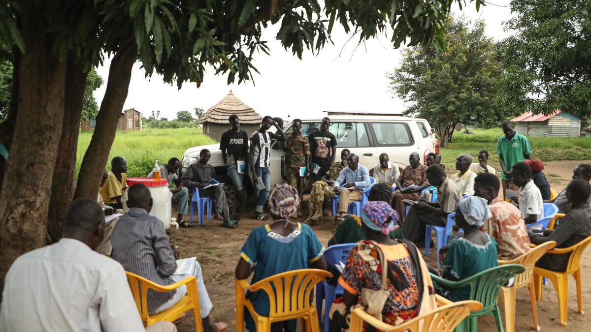 Community meeting held outdoors under a tree, with people seated in a circle on plastic chairs and several individuals standing near a white vehicle in the background.