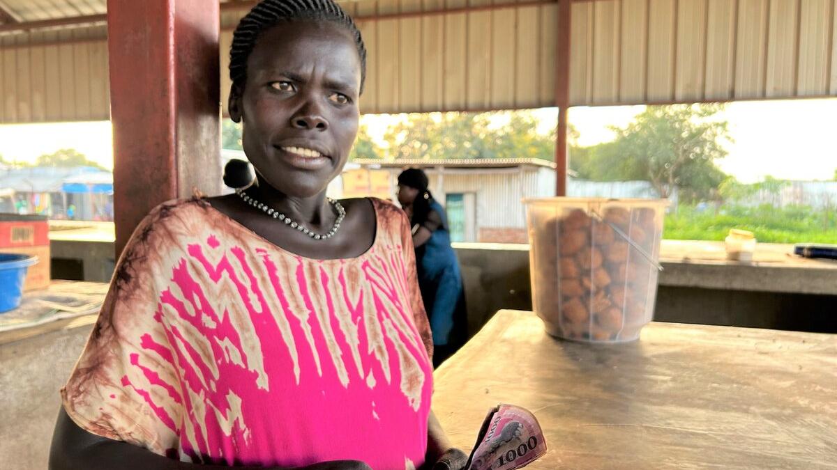 Person standing at a market stall holding currency notes, with a container of fried snacks on the counter and open-air structures in the background.