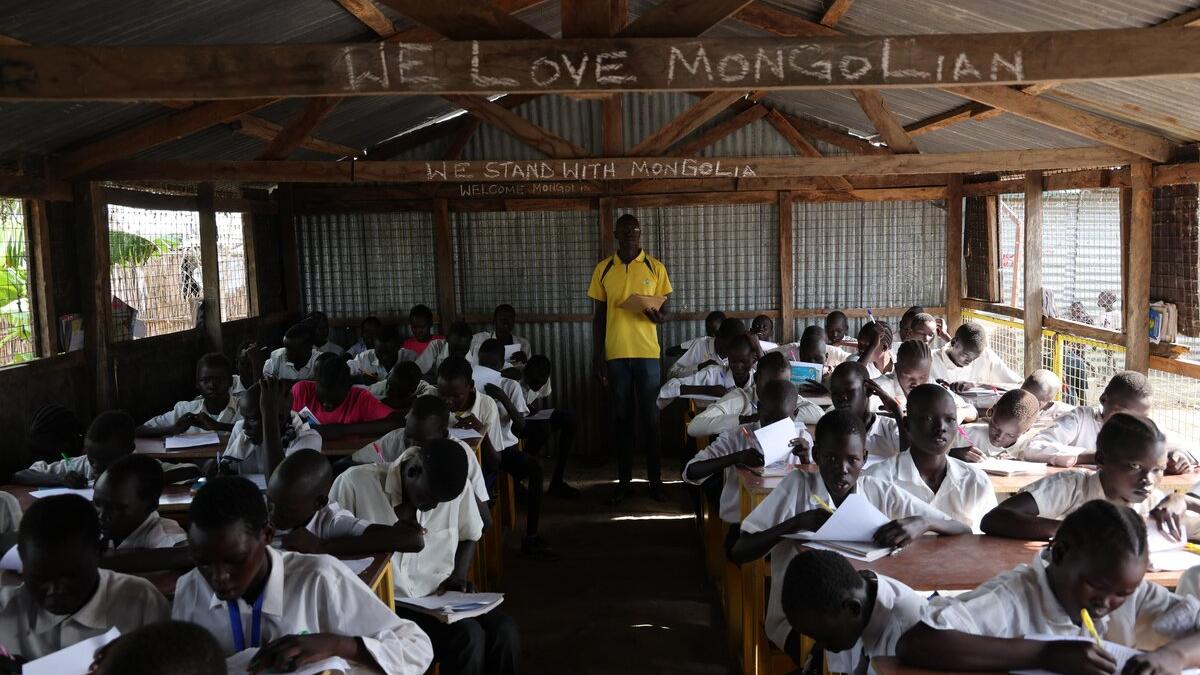A classroom with students in white uniforms seated at wooden desks, writing in notebooks, while an instructor stands at the front under a corrugated metal roof with chalk writing above.