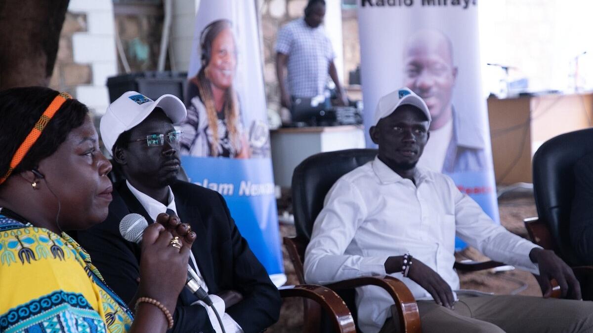 Three people seated indoors, one holding a microphone, with banners and a person standing near audio equipment in the background.