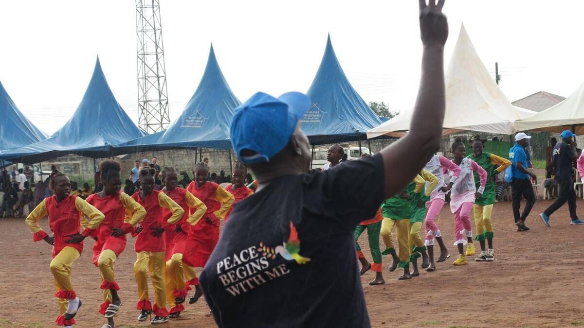 Outdoor event with a person wearing a shirt that reads “Peace Begins With Me” raising a hand, while groups of performers in colorful outfits dance in formation near blue tents.