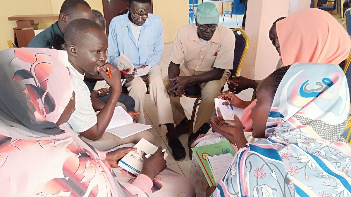 A group of people standing outdoors on a grassy field, some holding hands and others raising arms, appearing to participate in a group activity or celebration. Footwear is scattered on the ground, and a partly cloudy sky is visible in the background.