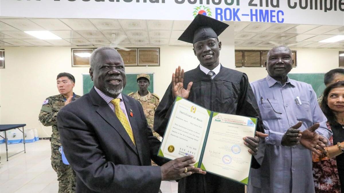 Graduation ceremony indoors with a person in a cap and gown holding a certificate, surrounded by others, with a banner for a vocational school visible above.