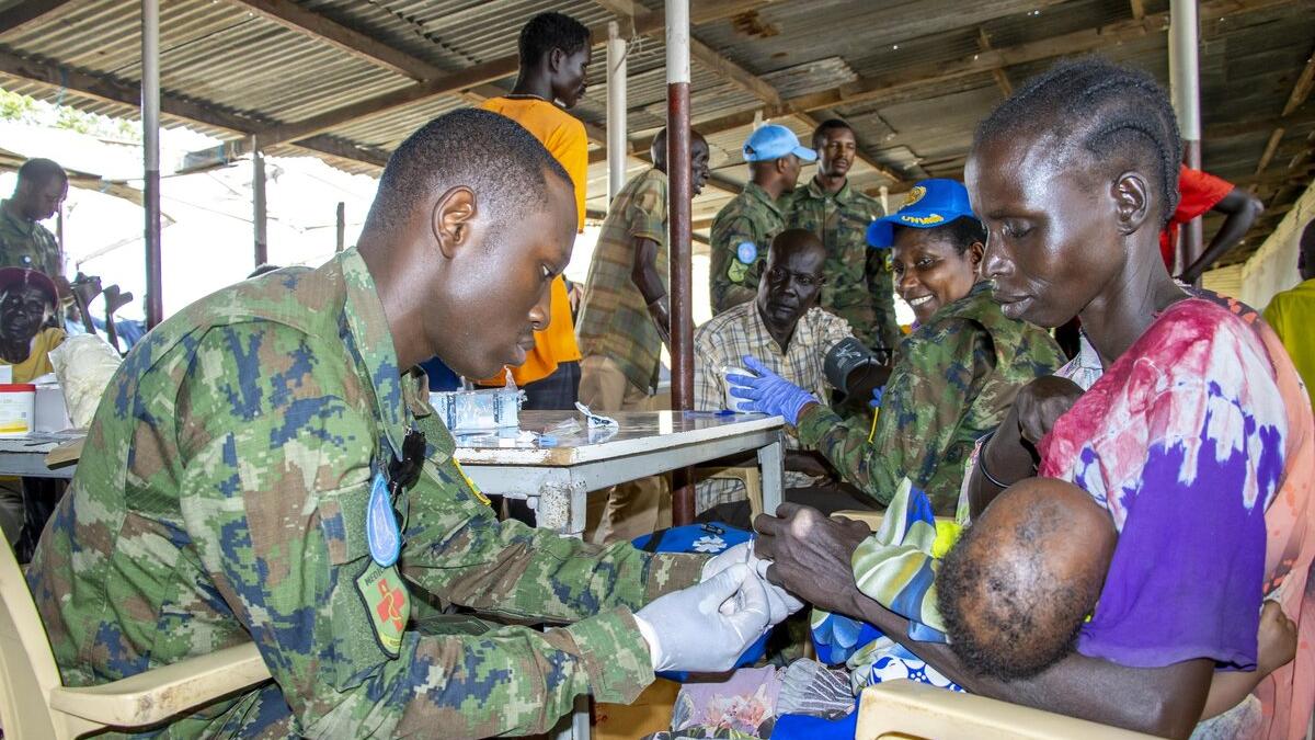 A person in camouflage uniform provides medical assistance to another person holding a baby at an outdoor clinic. Several people in uniforms and civilian clothing are seated at tables in the background under a corrugated metal roof.