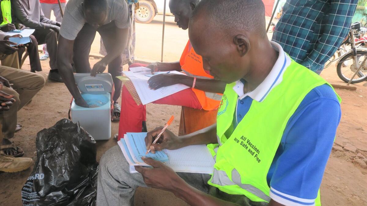 Health worker wearing a bright vest writes in a register during a vaccination campaign, with a cooler box and other people seated nearby.