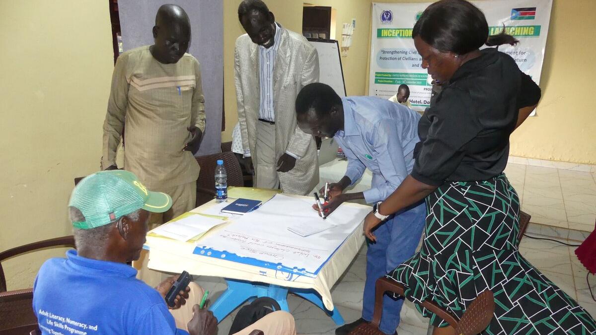 Group of people gathered around a table indoors, reviewing and writing on large sheets of paper during a workshop or planning session.