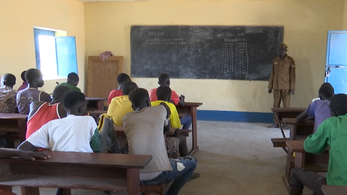 Classroom scene with students seated at wooden desks facing a chalkboard, where mathematical calculations and notes are written. An instructor stands near the board in front of the class.