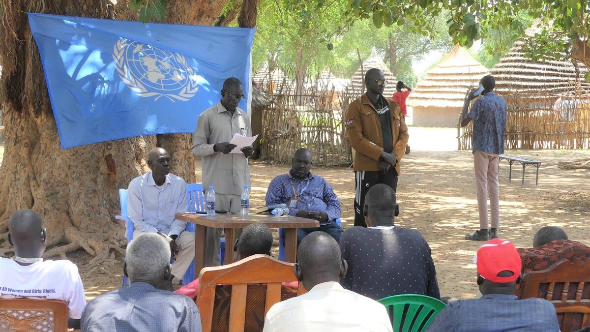 Outdoor meeting under a large tree with a UN flag displayed. Several people are seated on chairs facing a small table where others are standing and speaking. Traditional huts are visible in the background.