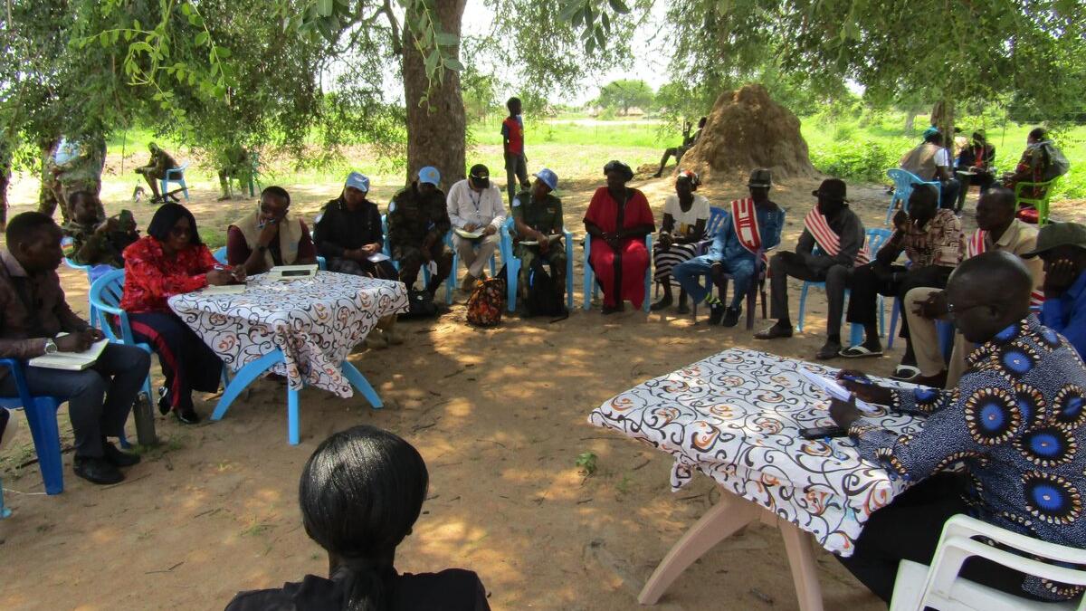 Group of people seated in a circle outdoors under a large tree, with two tables covered in patterned cloth at the center. Some participants wear blue caps, and traditional huts are visible in the background.