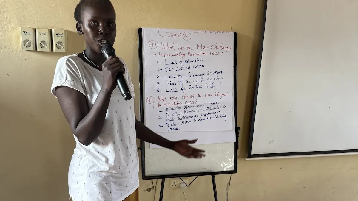 Person standing indoors holding a microphone and pointing toward a flip chart with handwritten notes about challenges in implementing Resolution 1325 and roles played. A blank whiteboard is positioned beside the flip chart.