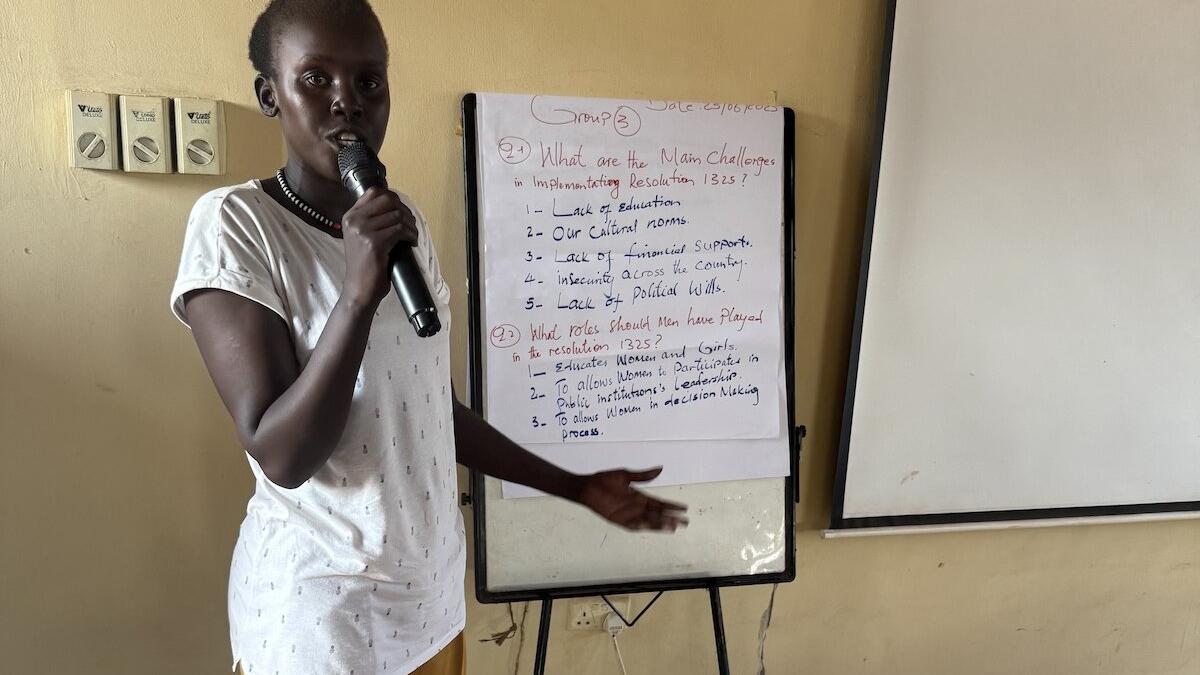 Person standing indoors holding a microphone and pointing toward a flip chart with handwritten notes about challenges in implementing Resolution 1325 and roles played. A blank whiteboard is positioned beside the flip chart.