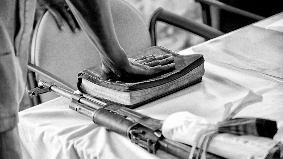 Close-up of a person’s hand resting on a worn book on a table, with a rifle placed beside it.