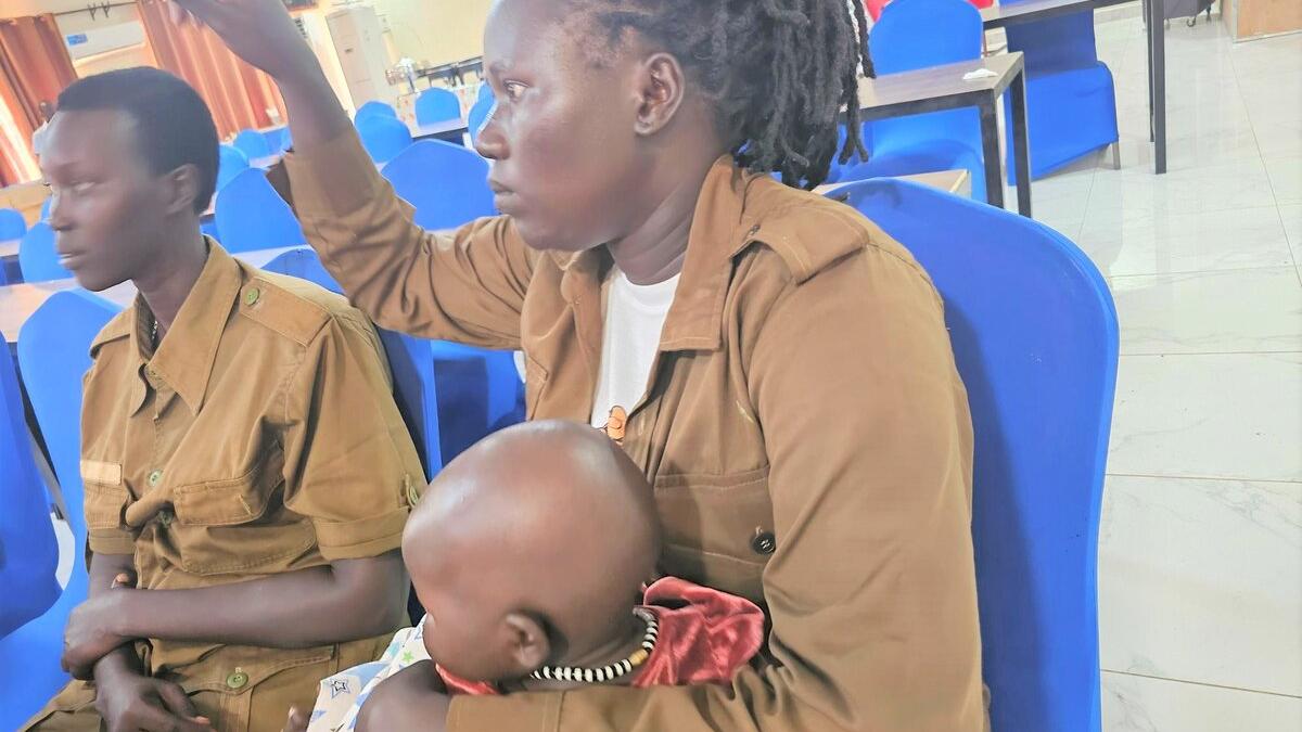 Two people in brown uniforms sit on blue chairs indoors, one holding a baby dressed in red, with tables and more chairs visible in the background