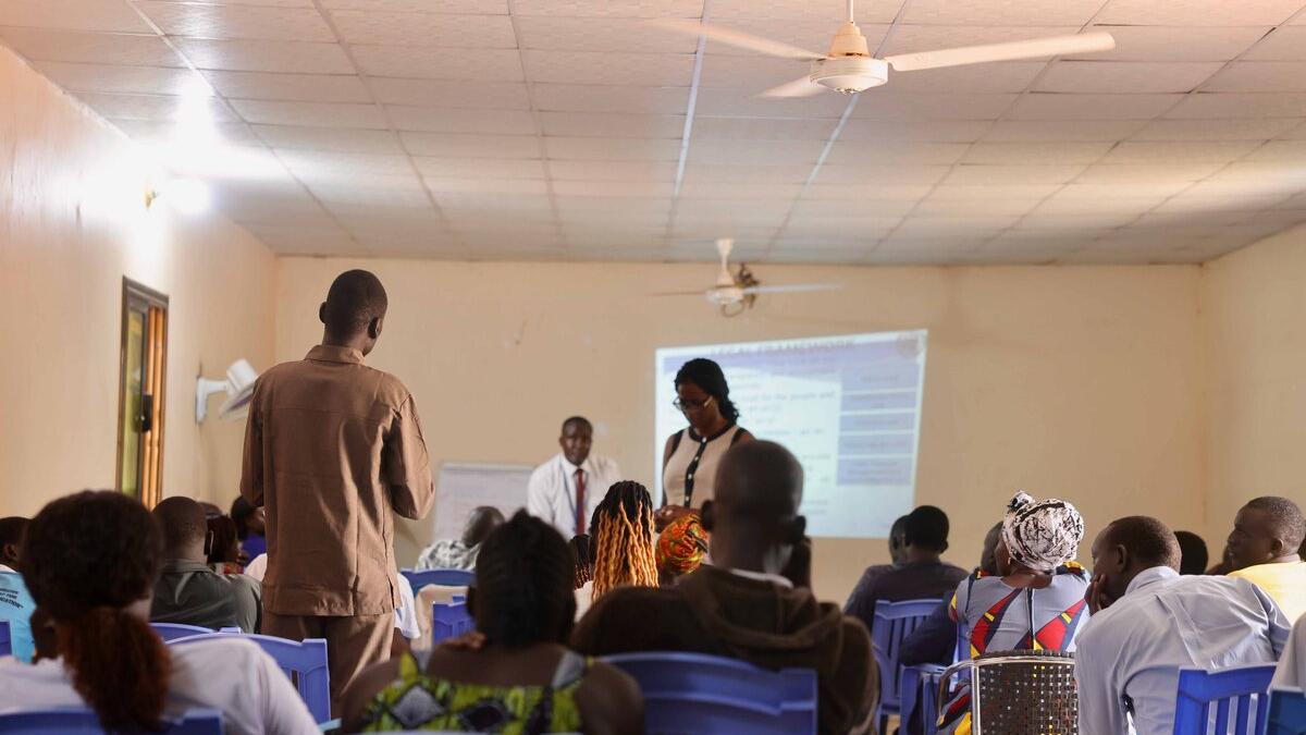 A classroom setting with people seated on blue chairs facing a projector screen, while two presenters stand at the front and one participant stands to speak.