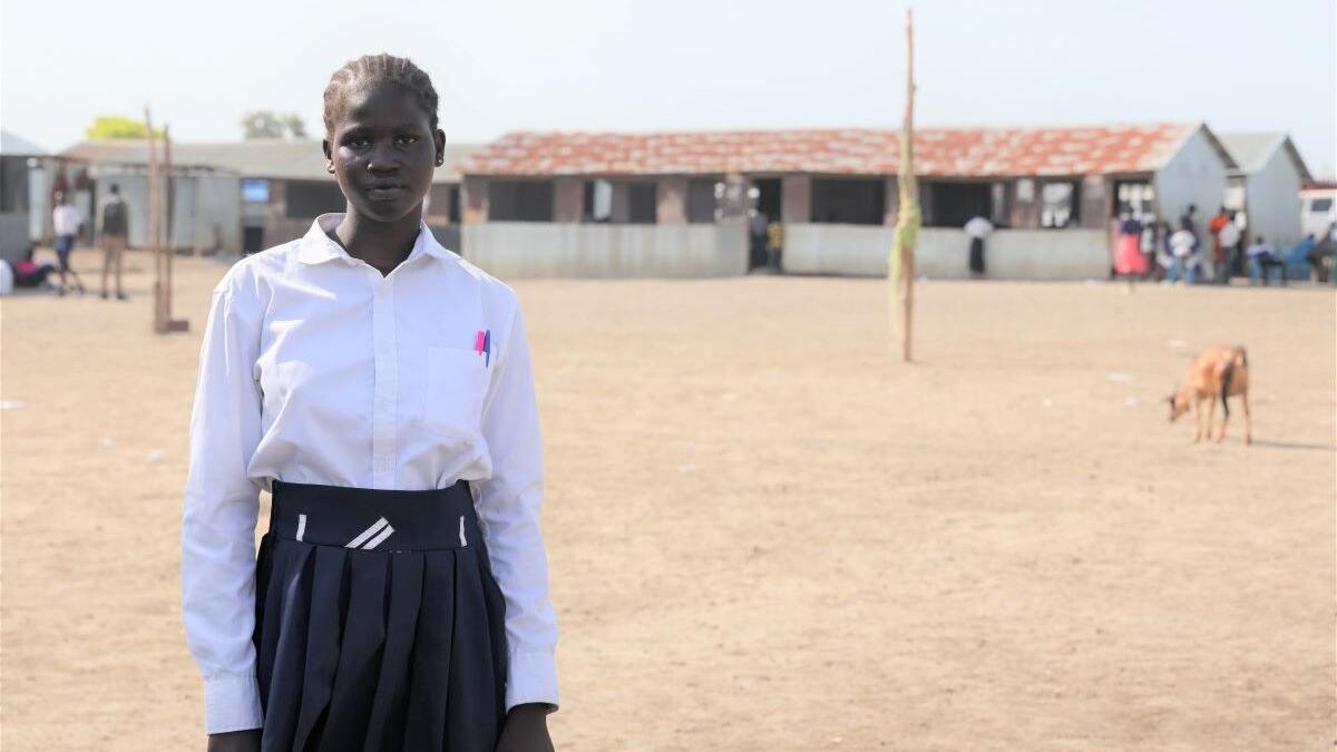 A student wearing a white shirt and dark pleated skirt stands on a dirt schoolyard with classrooms in the background and a goat grazing nearby
