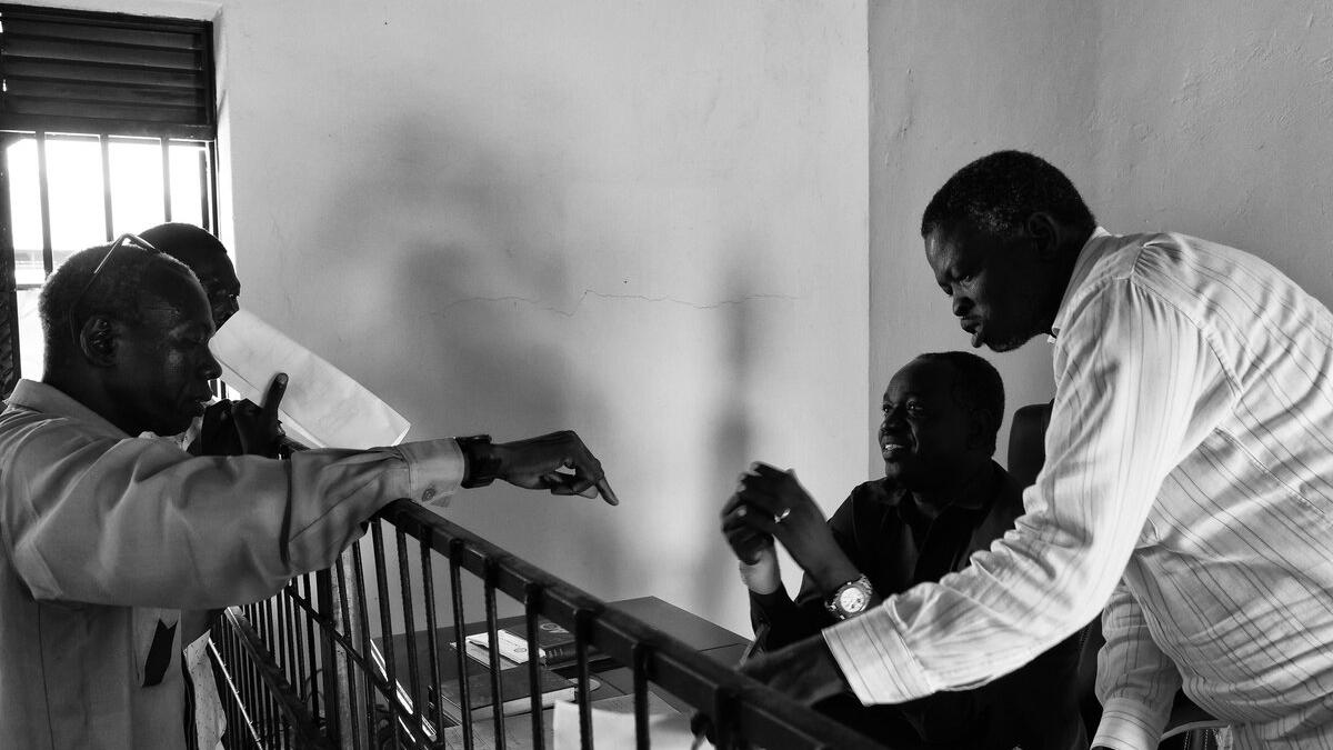 Three people engaged in discussion across a metal barrier inside a room with a barred window, papers and a notebook visible on the desk