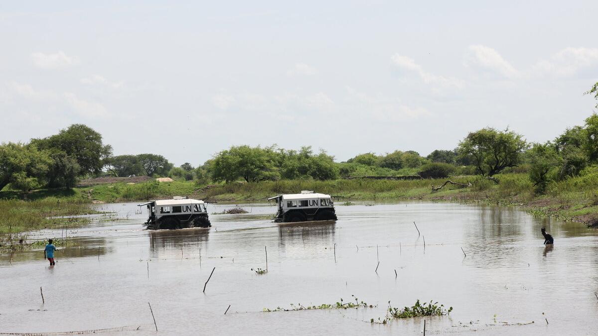 Two UN-marked armored vehicles driving through a shallow water body, with trees and vegetation in the background and two people standing in the water nearby.