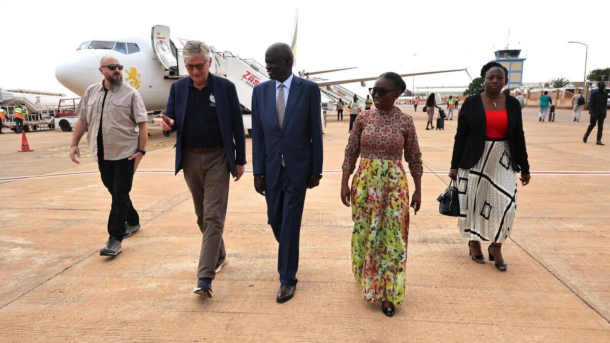 Four people including Under-Secretary-General Jean-Pierre Lacroix walking on an airport tarmac near a parked airplane, with other travelers and airport staff visible in the background.