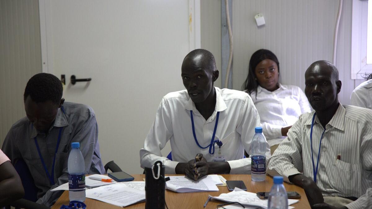 Several people seated at a table indoors during a meeting, with notebooks, papers, and water bottles spread across the table.