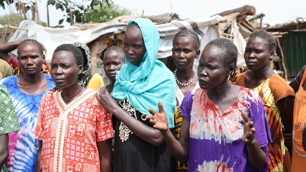 A group of people wearing colorful patterned clothing stand outdoors in front of makeshift shelters, with one person gesturing while others listen.