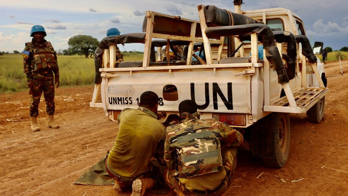 Two uniformed personnel crouch near the rear of a UN-marked pickup truck on a dirt road, while another stands nearby wearing a blue helmet.