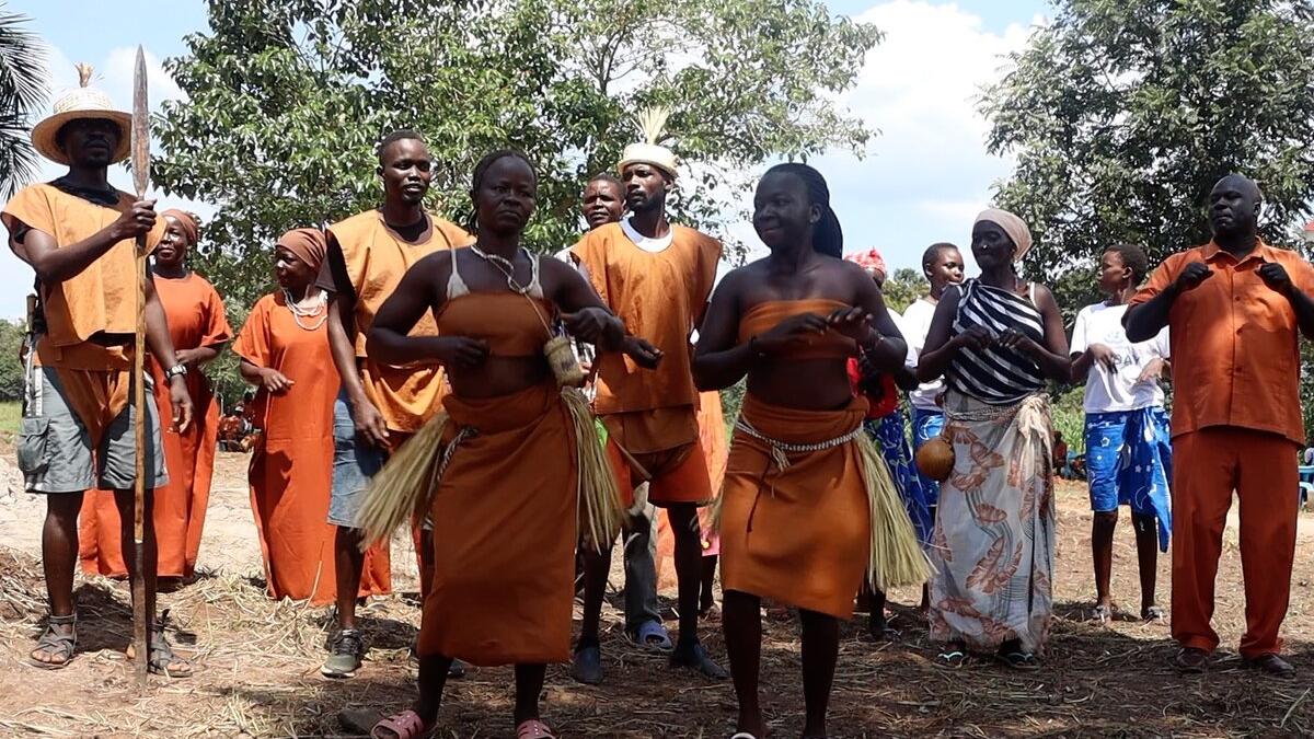 Group of people outdoors wearing traditional orange attire, some with grass skirts and necklaces, standing together with trees in the background.