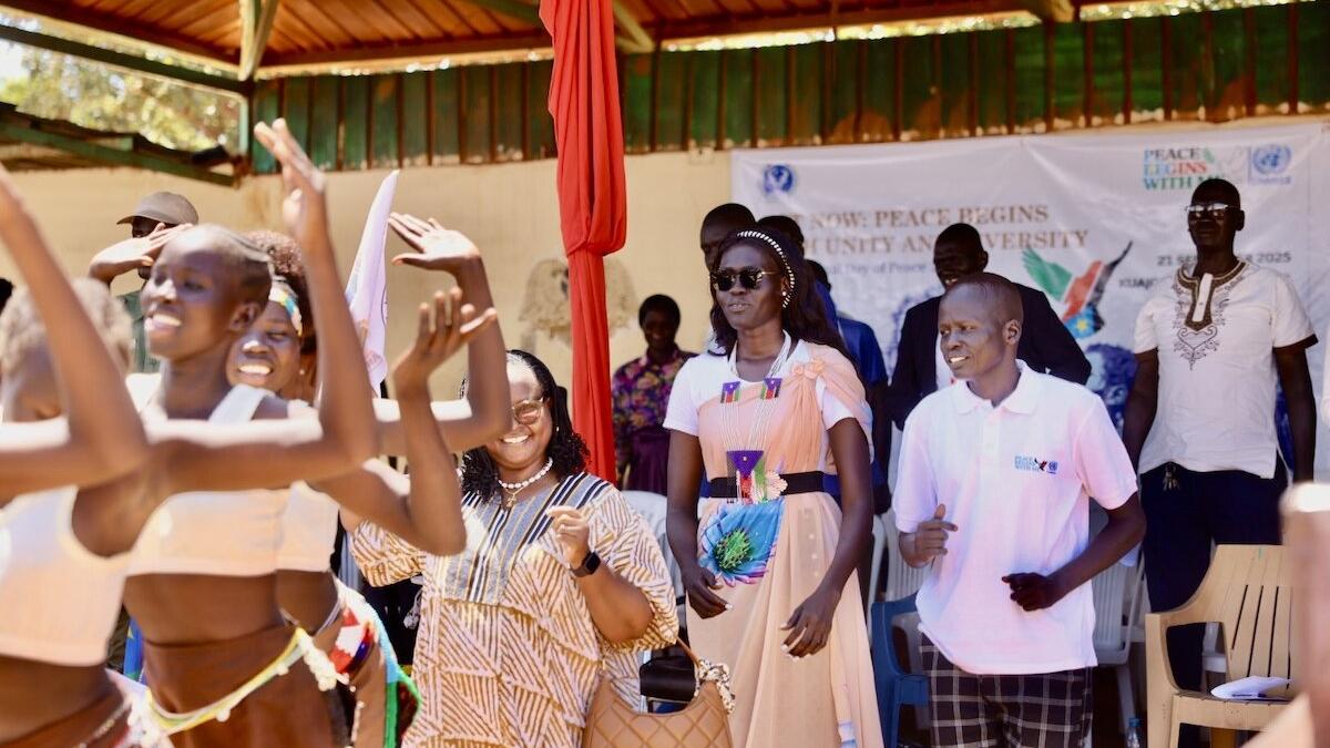 People standing and dancing under a covered outdoor structure during an event, with a banner in the background displaying the words ‘Peace Begins’ and other text.