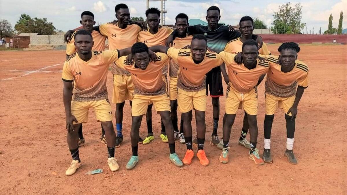 A soccer team wearing matching yellow and black uniforms poses together on a dirt field, with goalposts and trees visible in the background