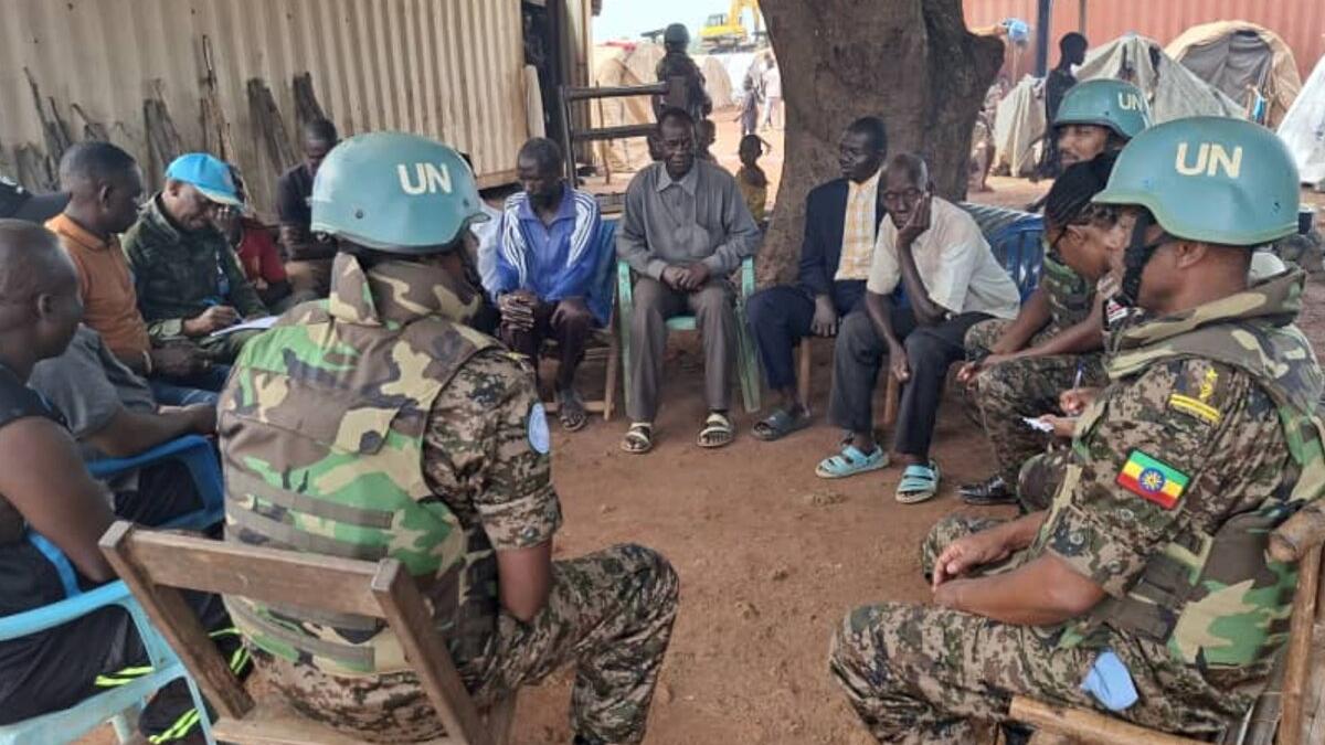 UN peacekeepers in camouflage uniforms and blue helmets sit in a circle outdoors, engaging in discussion with local community members near tents and a corrugated structure