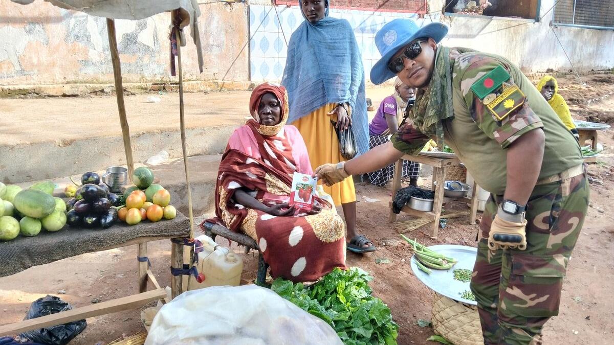 A person in camouflage uniform hands a leaflet to a seated vendor at an outdoor market stall with fruits and vegetables displayed on a mat