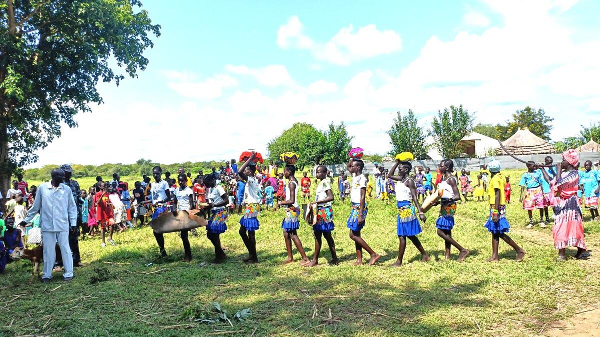 A group of children in colorful clothing and blue skirts perform a traditional dance in a grassy field, with a crowd of spectators and trees in the background.