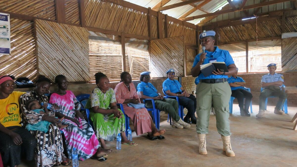 A UN peacekeeper in blue uniform stands and speaks with a notebook and microphone during a community meeting inside a wooden structure, while others sit on blue chairs listening