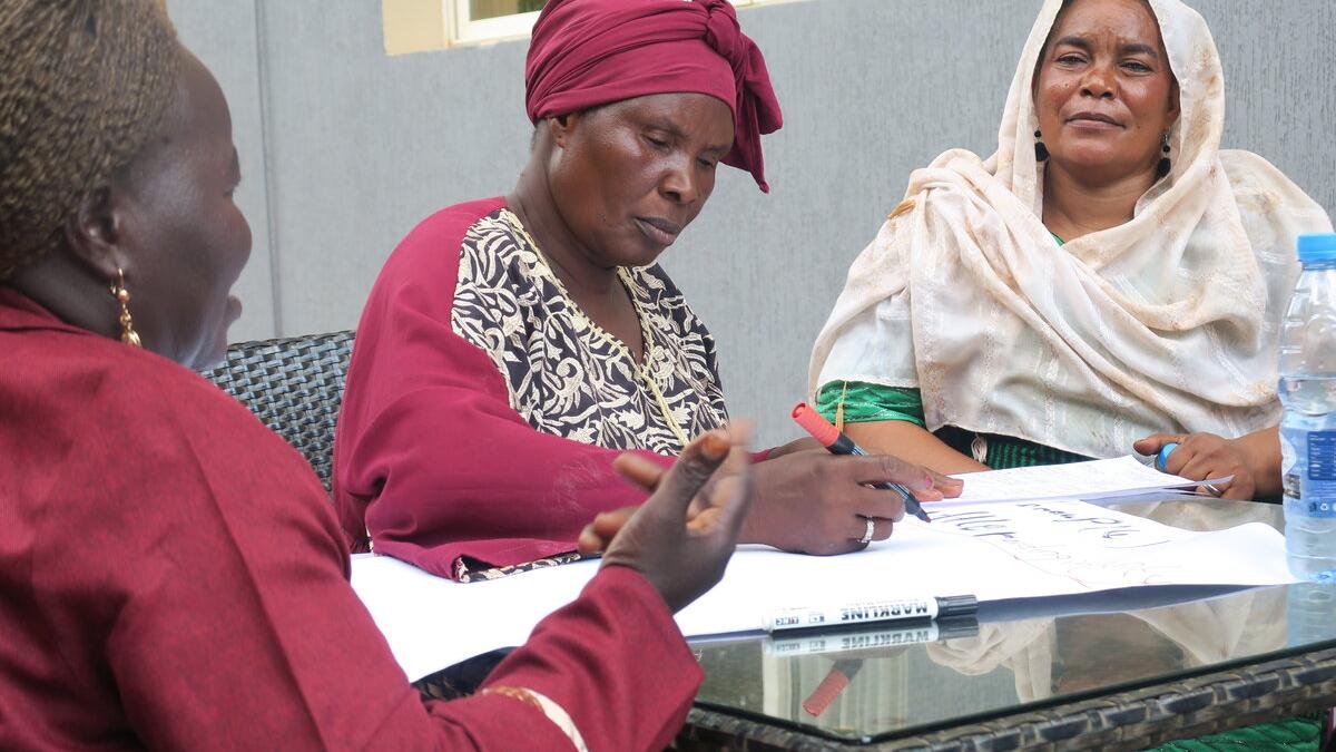 A community meeting in a room with orange plastic chairs arranged in rows, participants seated and listening to a speaker at the front near posters and a banner on the wall.
