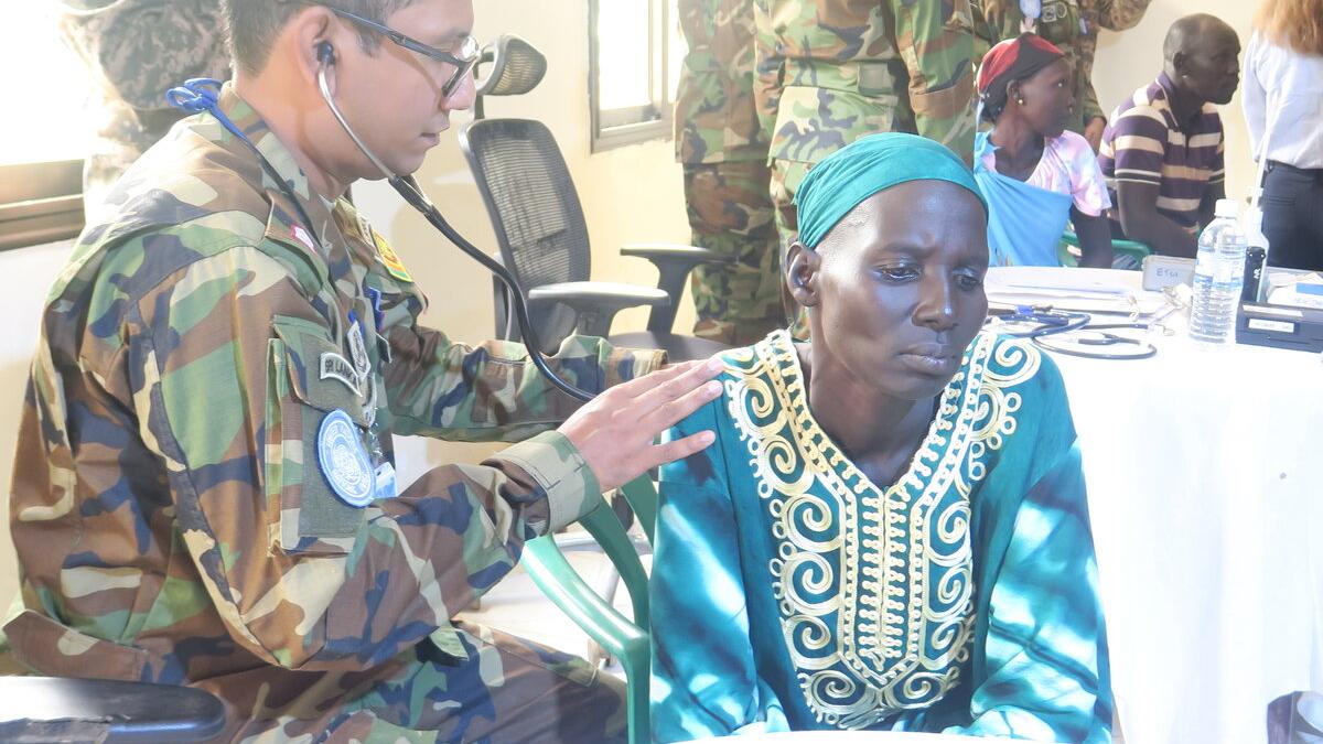 A person in camouflage uniform uses a stethoscope to examine another person wearing a turquoise garment with intricate embroidery during a medical check-up indoors