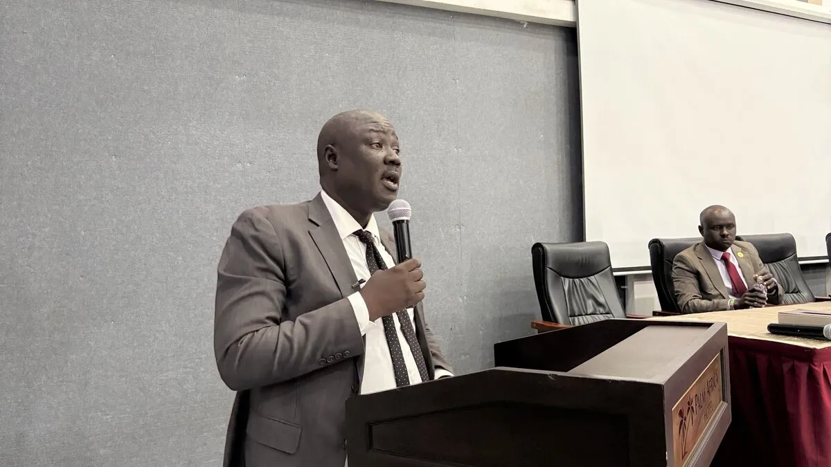 A person in a suit speaks at a podium with a microphone in a conference room, while another person sits at a table with documents and a microphone in front.