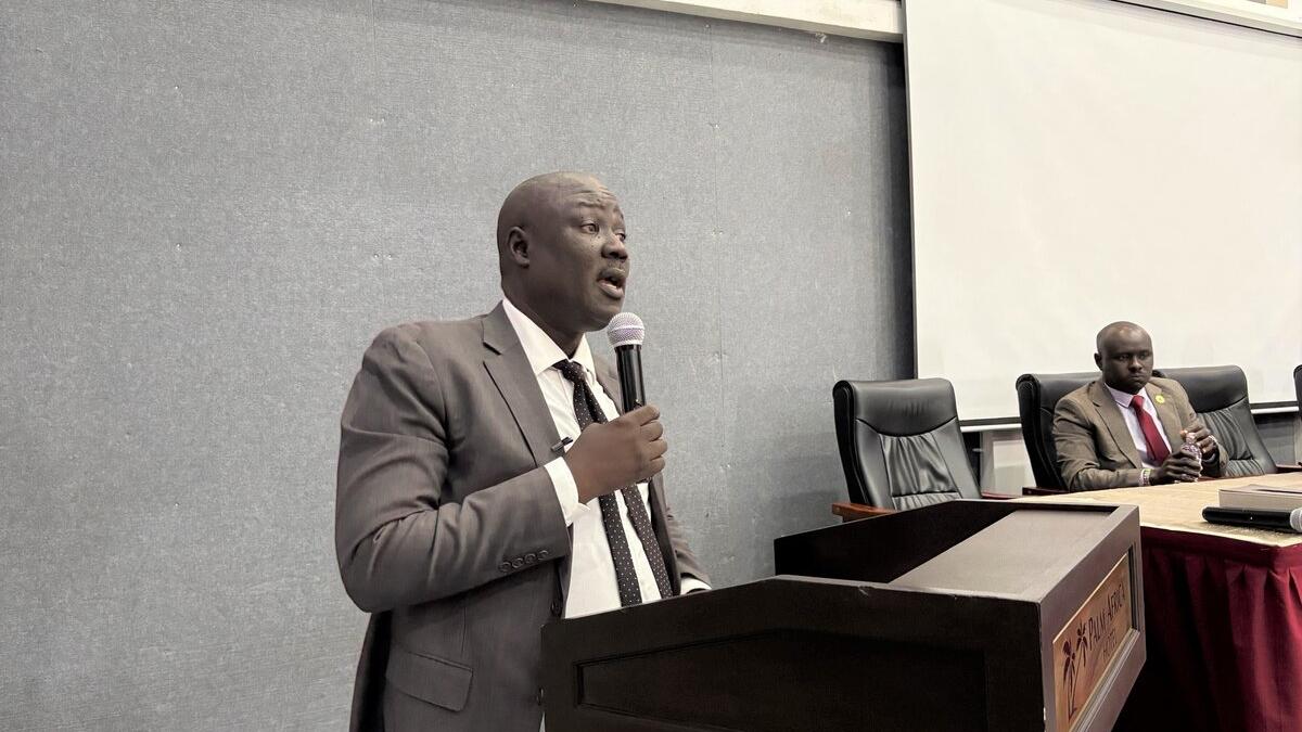 A person in a suit speaks at a podium with a microphone in a conference room, while another person sits at a table with documents and a microphone in front.